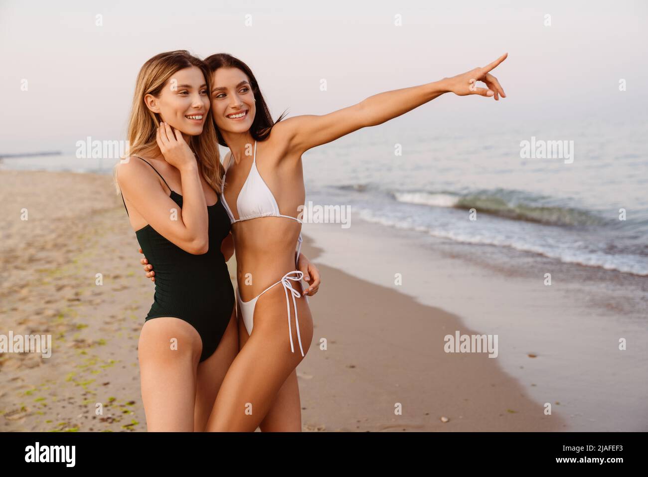 Young white women hugging and pointing finger aside while posing on beach Stock Photo