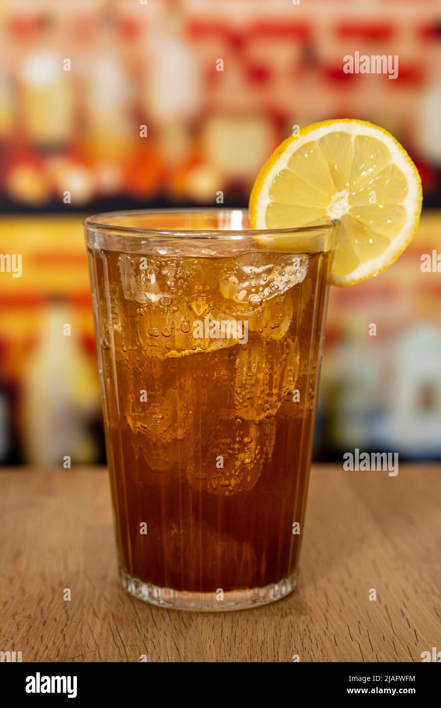 Glass with sparkling cola, ice and lemon on a wooden counter in front of a bar. Stock Photo