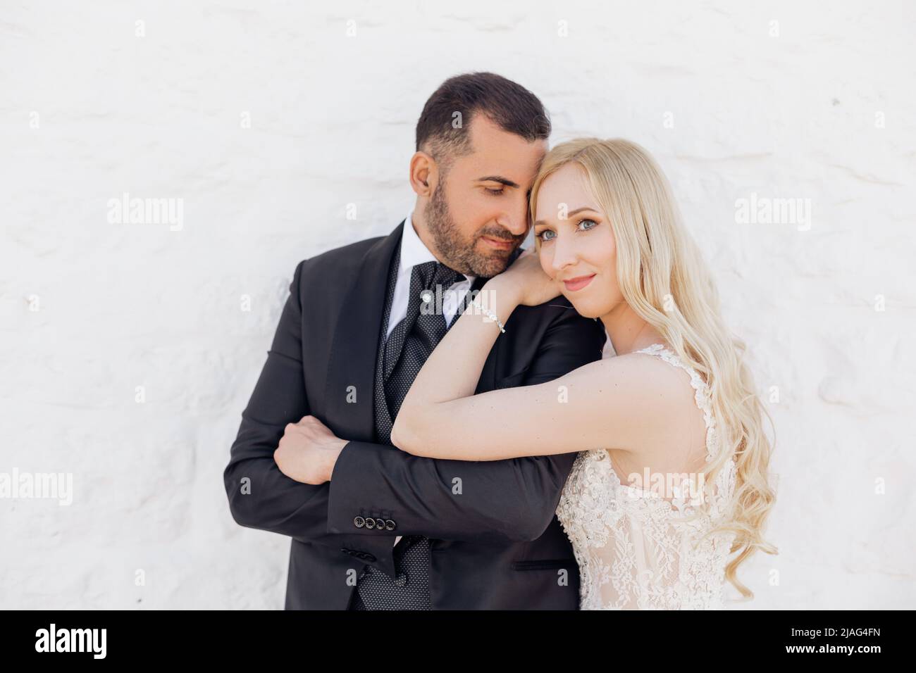 Portrait of young blonde bride in white dress and brunet groom in suit embracing and walking down street in Italy closeup, white wall background Stock Photo