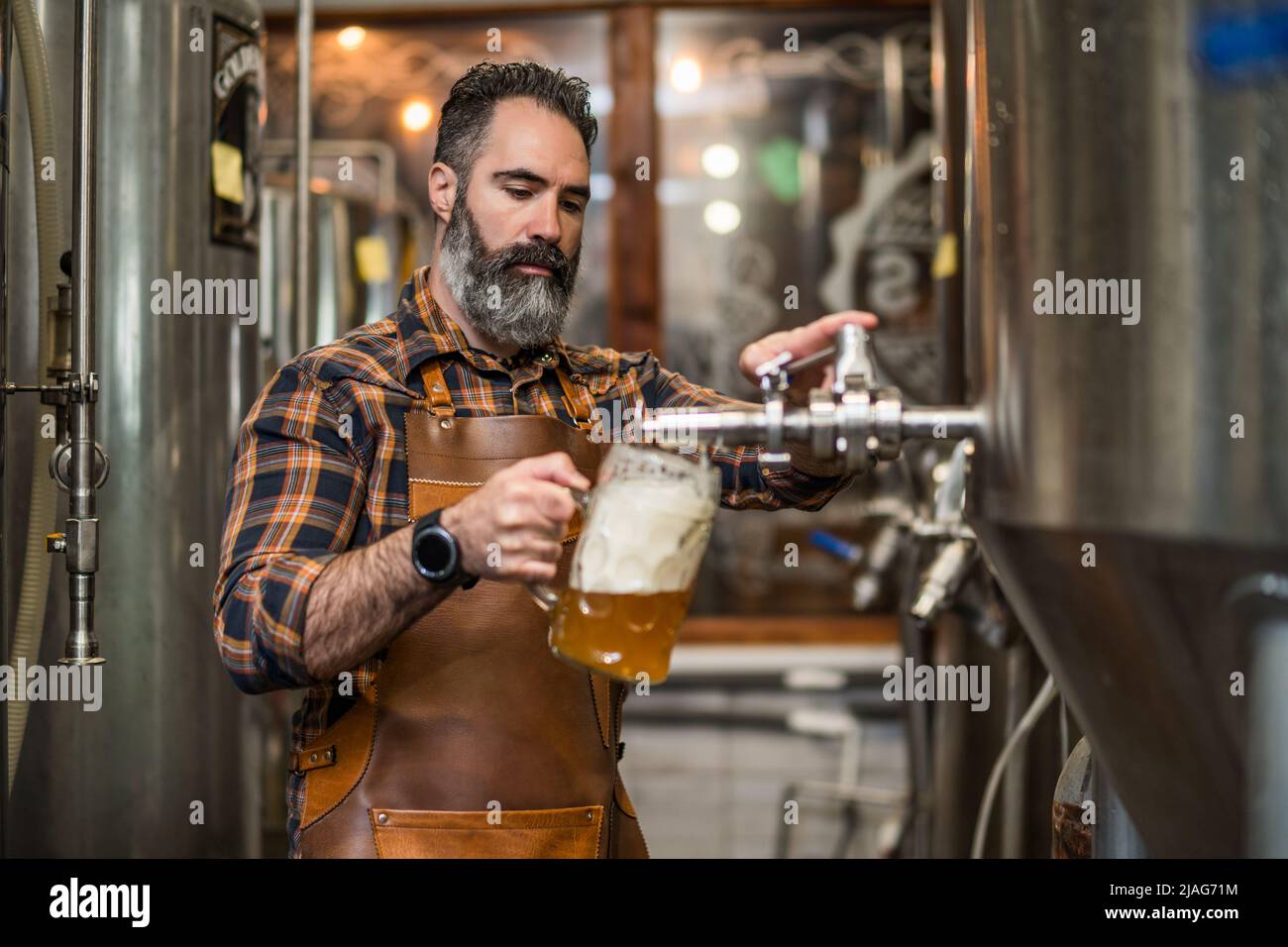 Bearded brewery master tasting beer in production facility. Small