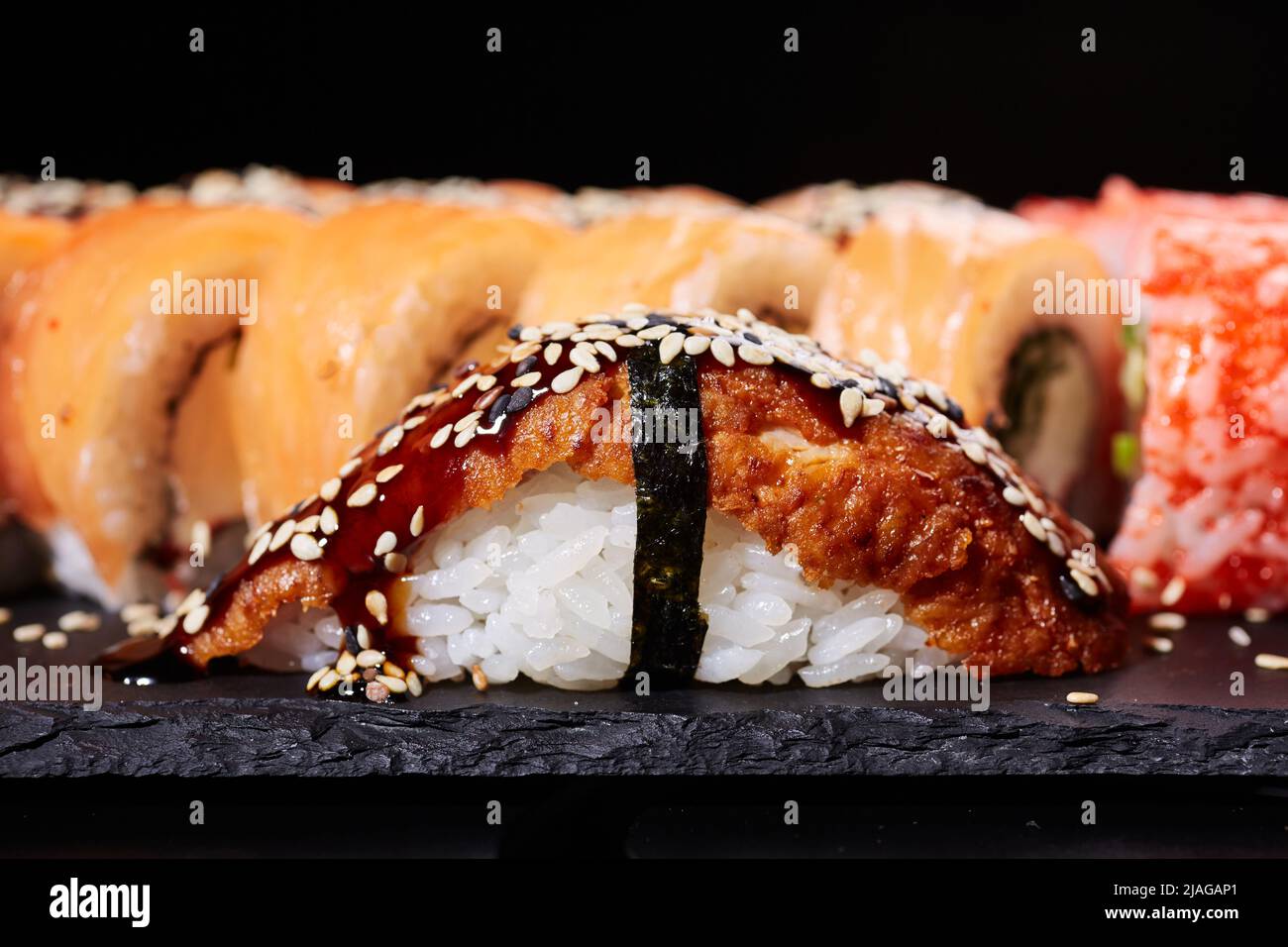 Close Up Of One Piece Of Nigiri Sushi With Smoked Salmon Soy Sauce And Sesame Seeds On A Background Of Other Sushi Served On A Slate Of Black Stone Stock Photo