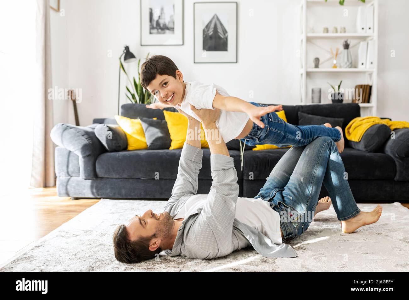 Happy father and school-age son spending time together at home, have fun lying on the floor in cozy living room, dad carrying boy, son spreading arms pretends airplane. Lovely father play with sonny Stock Photo