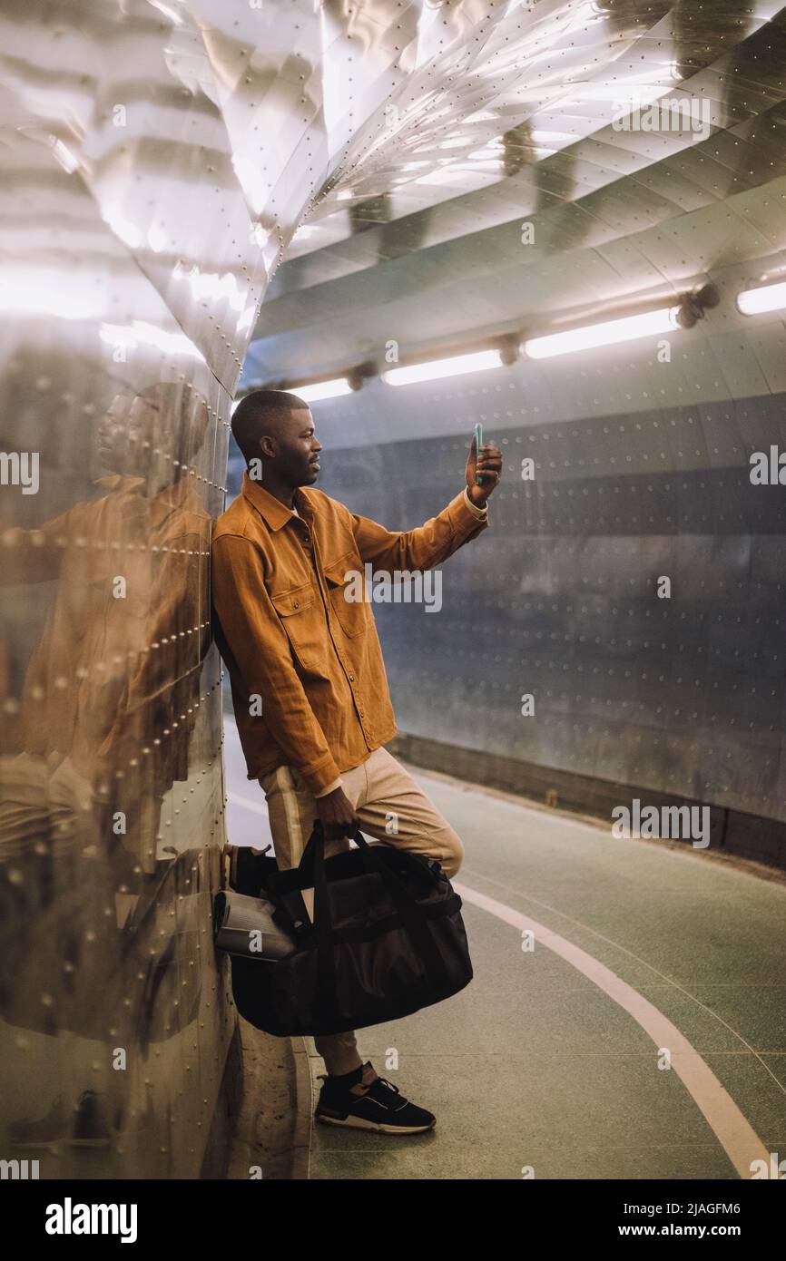 Full length of young man carrying bag while taking selfie through mobile phone in subway tunnel Stock Photo