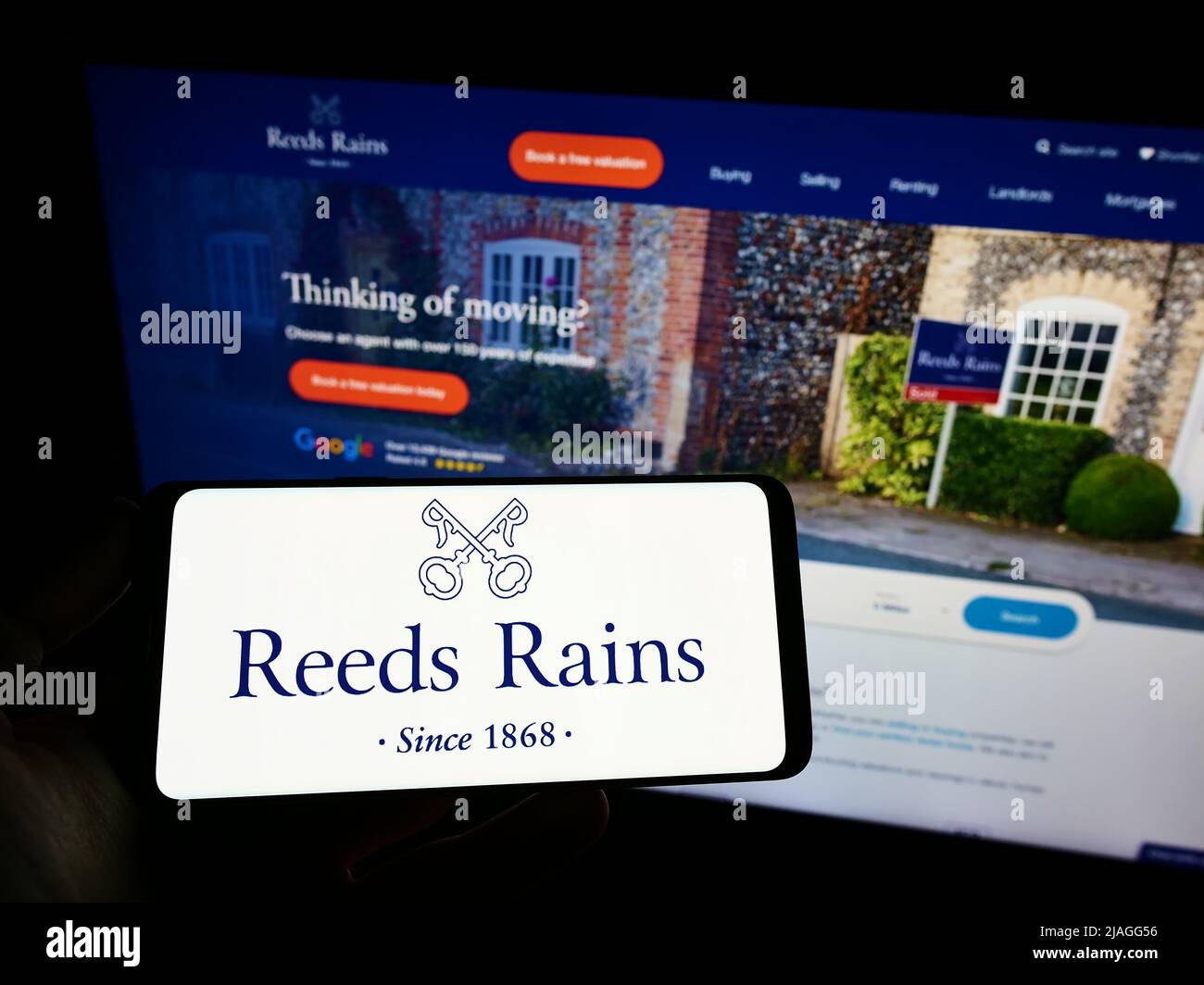 Person holding smartphone with logo of British real estate company Reeds Rains Limited on screen in front of website. Focus on phone display. Stock Photo