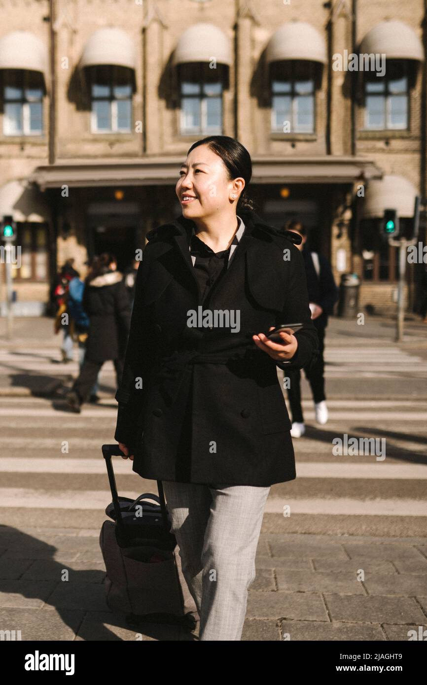 Smiling businesswoman with luggage holding smart phone while crossing street in city Stock Photo