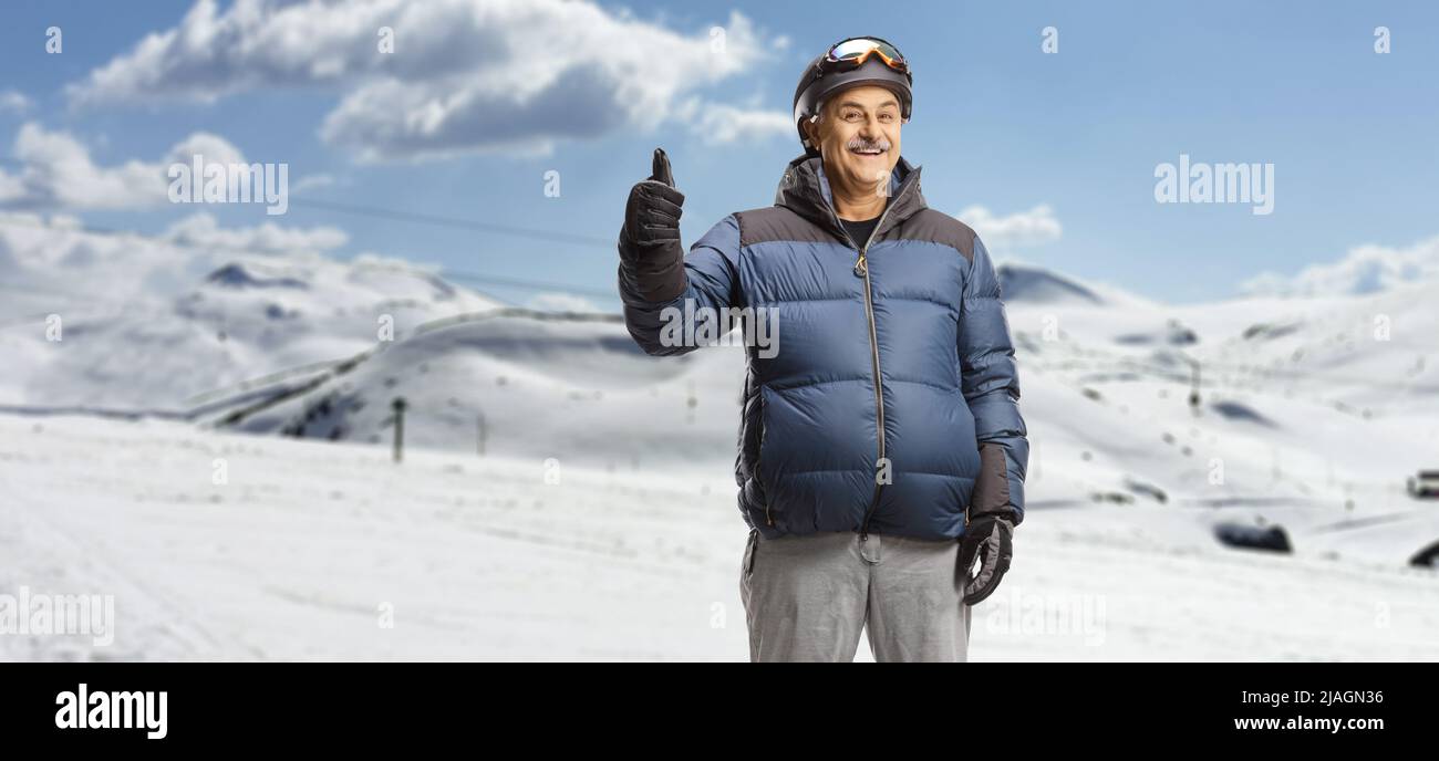 Mature man in a winter jacket standing in a ski resort on a snowy mountain and gesturing thumb up Stock Photo