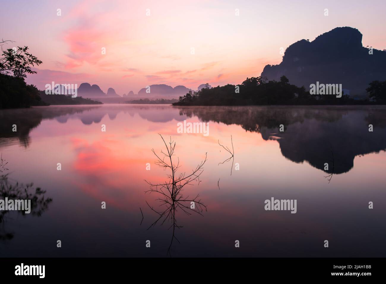 Landscape of the tropical lake during sunrise, dramatic clouds and sunrise sky reflection on the lake, morning mist covering mountains in background. Stock Photo