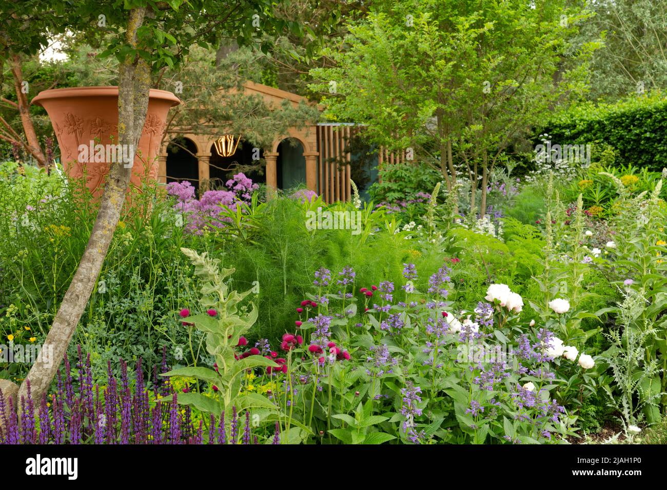 A large decorative urn and a classical style oak pavilion surrounded by lush herbaceous borders, trees and stone paving in the RNLI Garden designed by Stock Photo