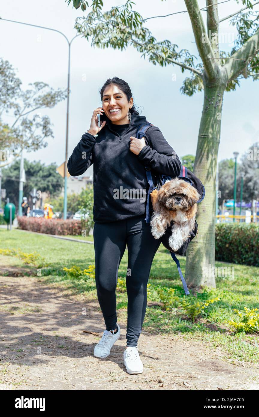 Young smiling woman carrying her dog in a backpack while talking on the phone, outdoor in nature Stock Photo