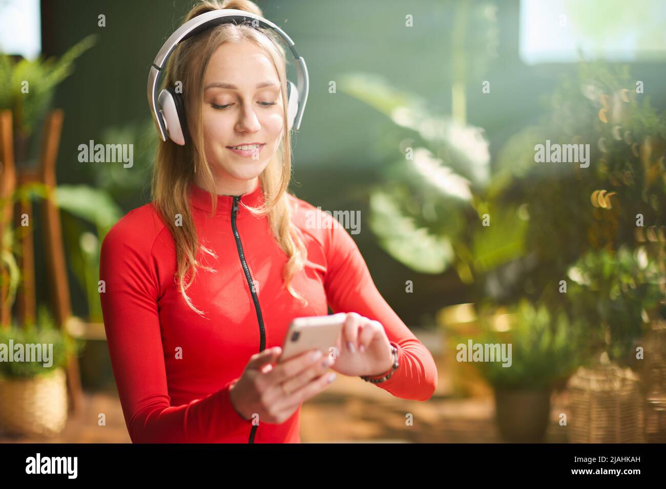 smiling young female in red fitness clothes with headphones using smartphone at modern green home. Stock Photo