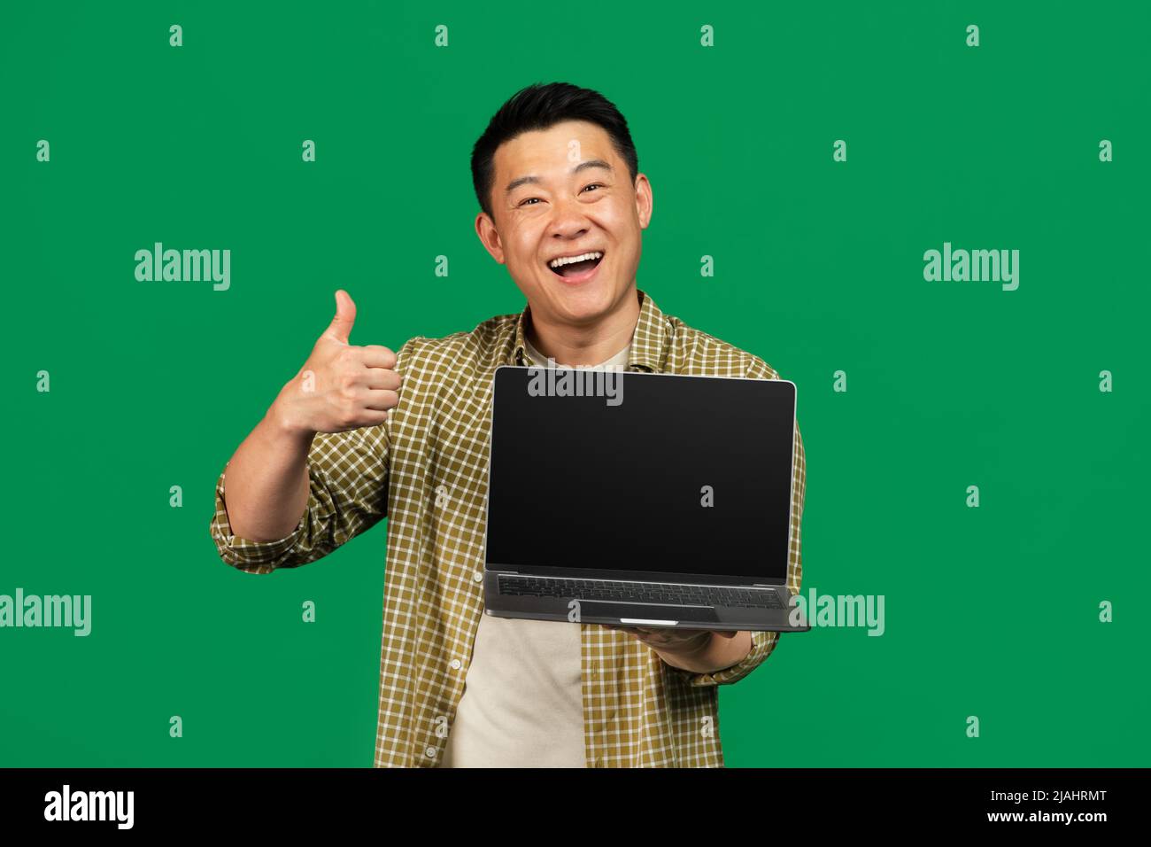 Happy asian mature man showing laptop empty screen and gesturing thumb up, posing over green background Stock Photo