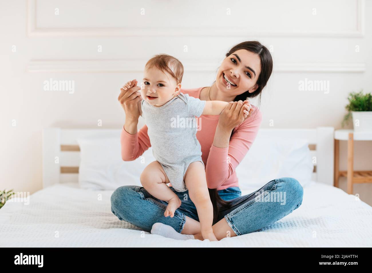 Happy young mom helping to her infant child making first steps, sitting on bed, spending time with little kid Stock Photo