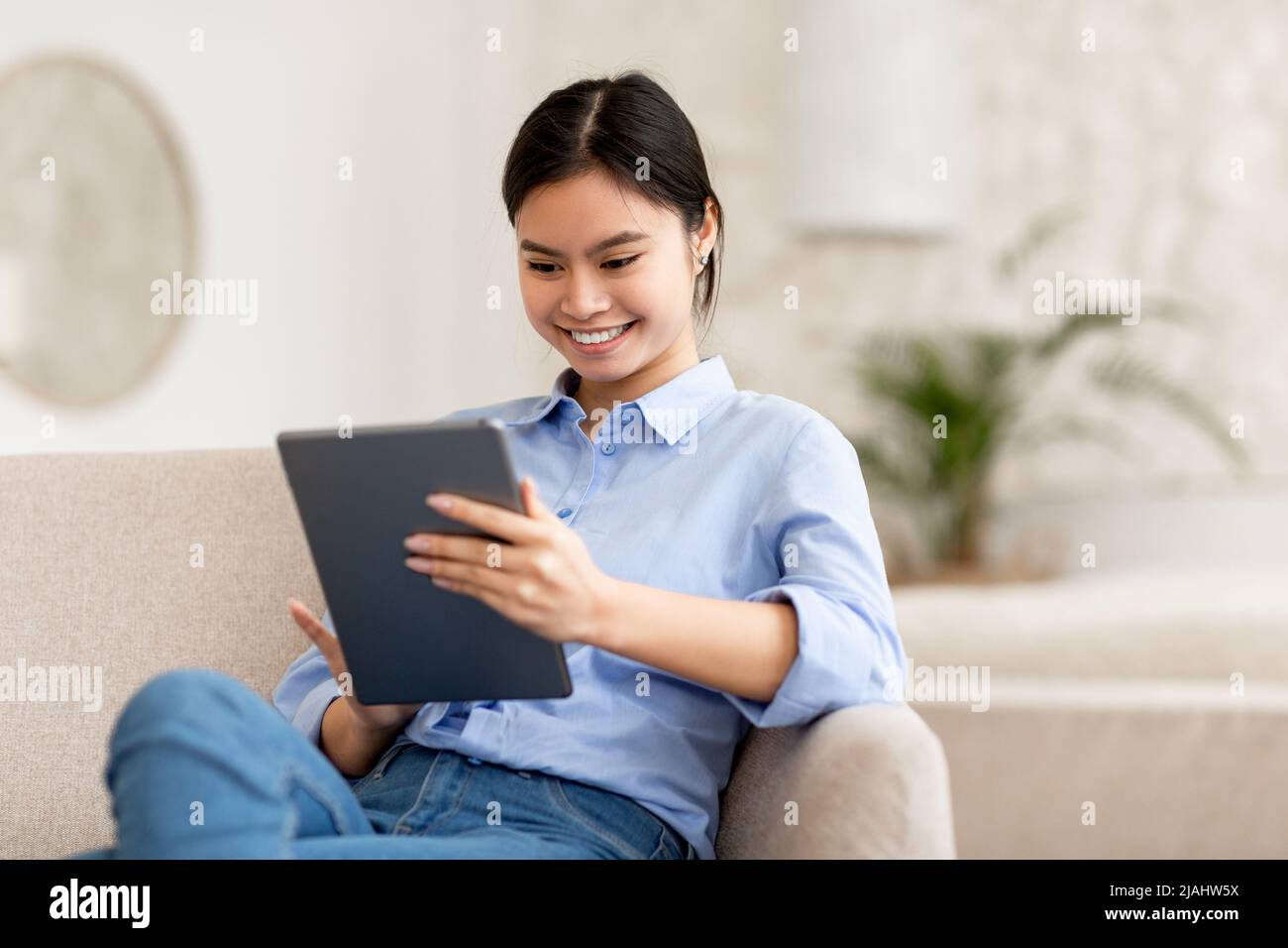 Smiling lady sitting on sofa, using digital tablet Stock Photo