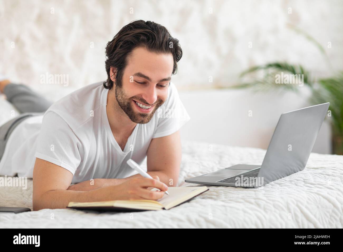 Smiling young man laying on bed, using laptop, taking notes Stock Photo