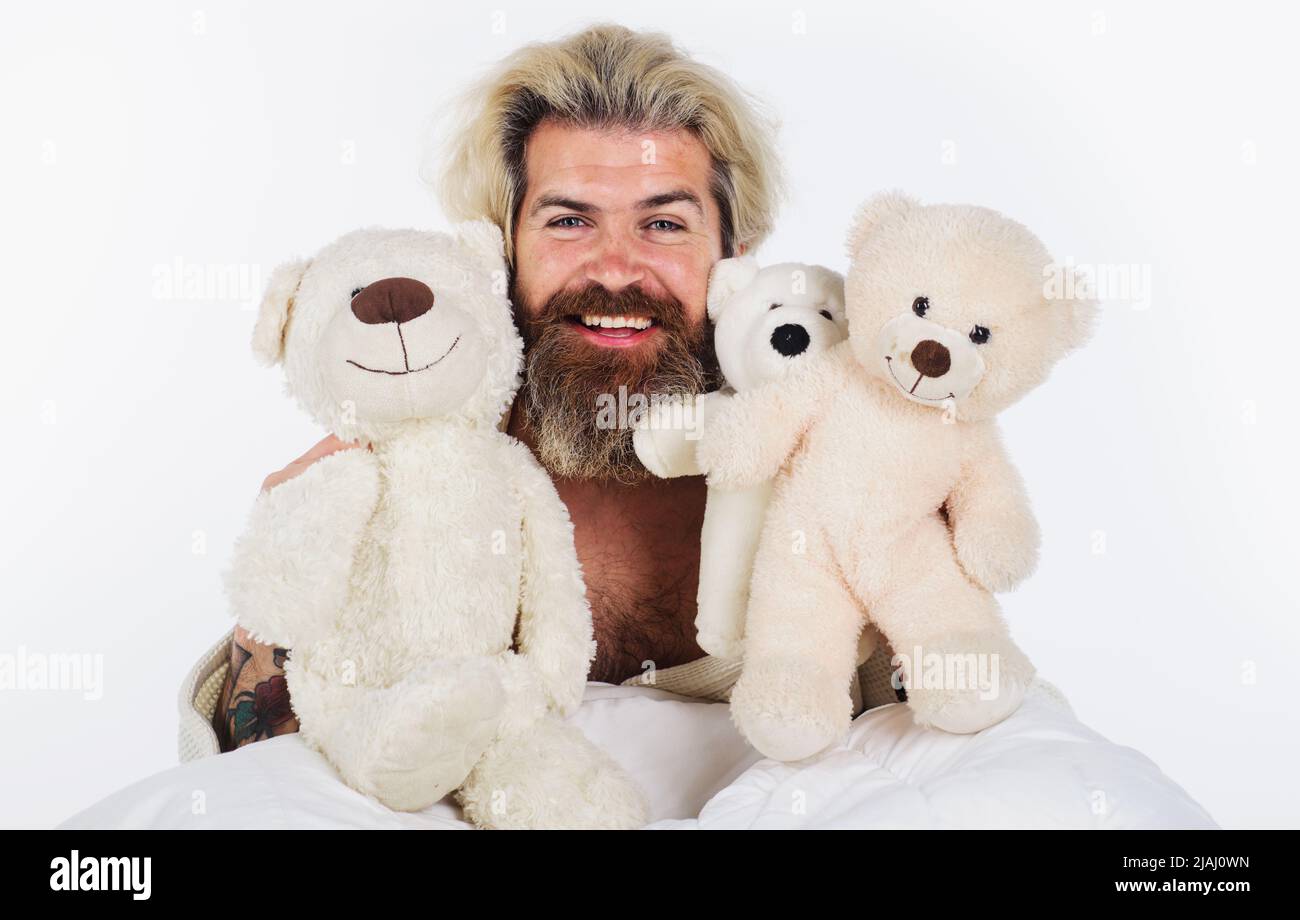 Smiling man with teddy bears in bed. Bearded guy with plush toys. Good morning. Sleeping toy. Stock Photo