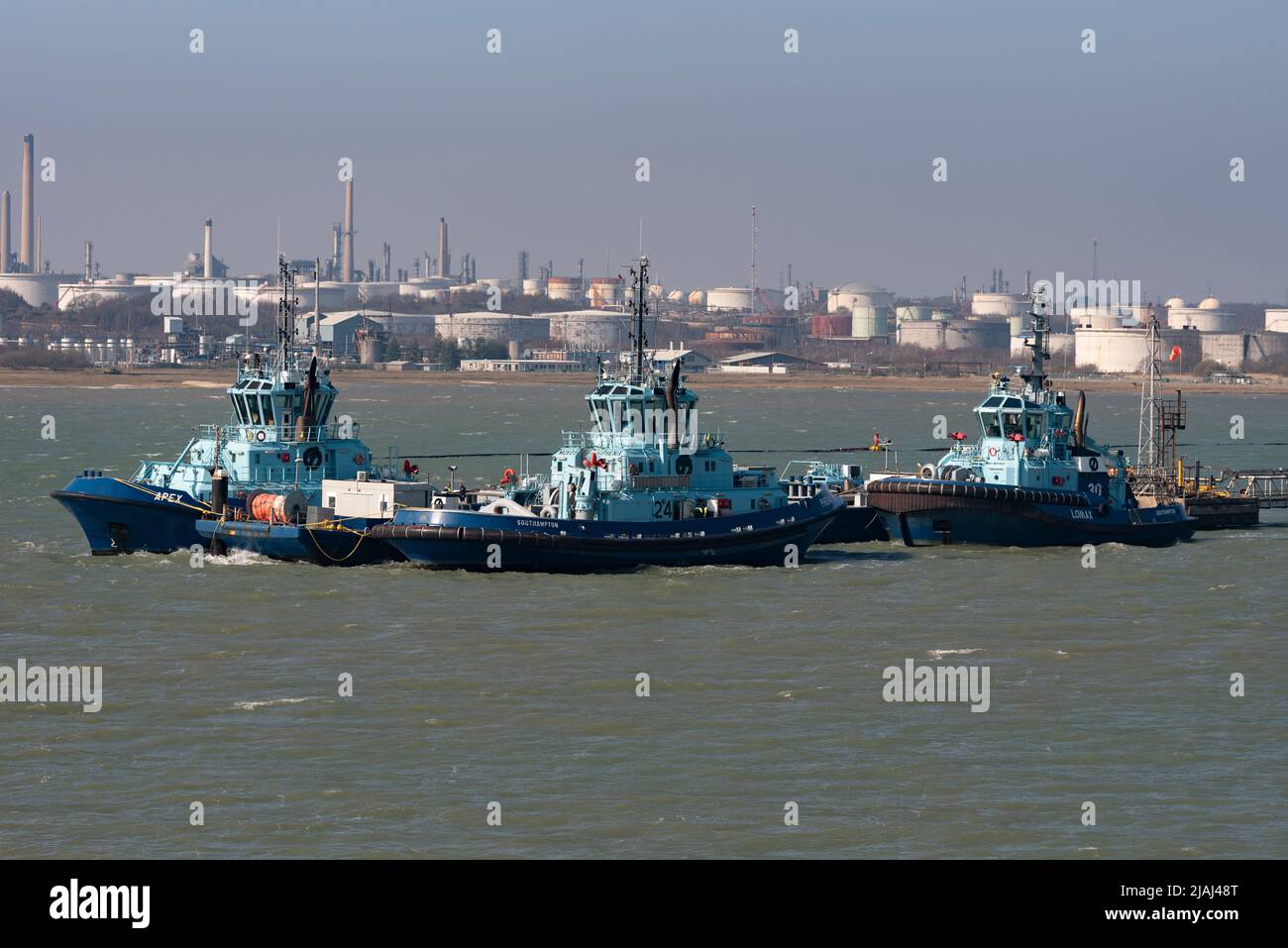 Fawley, Southampton, England, UK. 2022.  Three ocean going tugs alongside a jetty with a background of Fawley Refinery, chimneys and smoke stacks, Stock Photo
