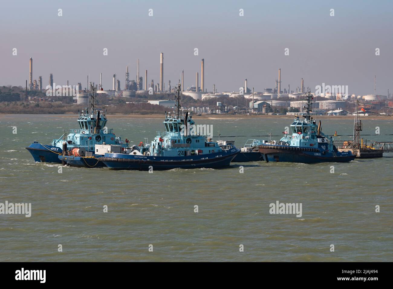 Fawley, Southampton, England, UK. 2022.  Three ocean going tugs alongside a jetty with a background of Fawley Refinery, storage tanks, chimneys and sm Stock Photo
