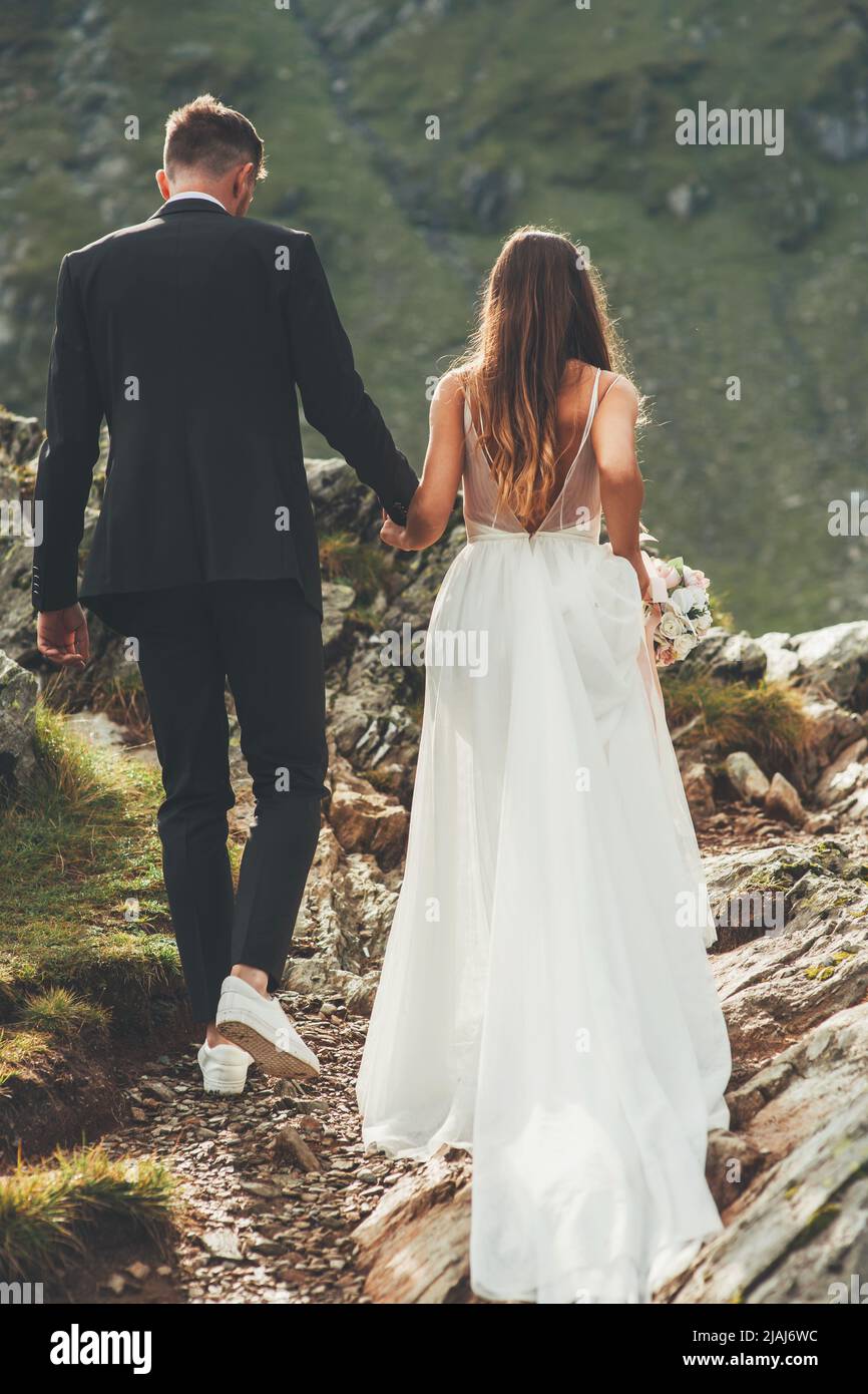 Back view of a groom and bride in a white dress walking, holding hands ...