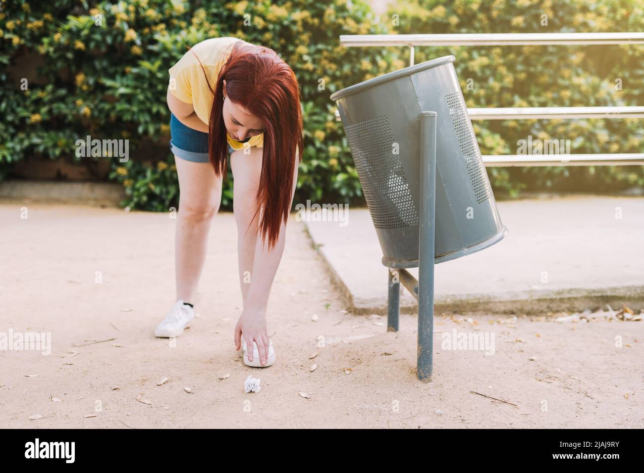 young woman bending down to pick up litter from the ground in a public park, to deposit it in a litter bin. young environmentalist in the city. Stock Photo