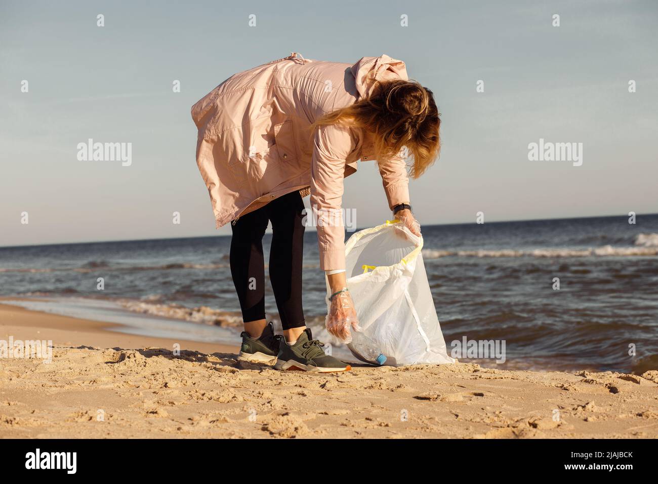 Woman volunteer and activist cleaning up sandy beach near sea from plastic waste, garbage. Picking up bottles to bag Stock Photo