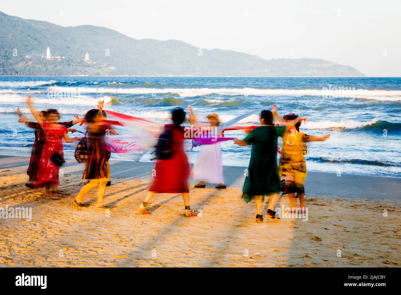 Motion blur of a group of Asian women dancing in a circle on the beach at Da nang, VN. Hua Linh Ung statue in background on mountain. Stock Photo