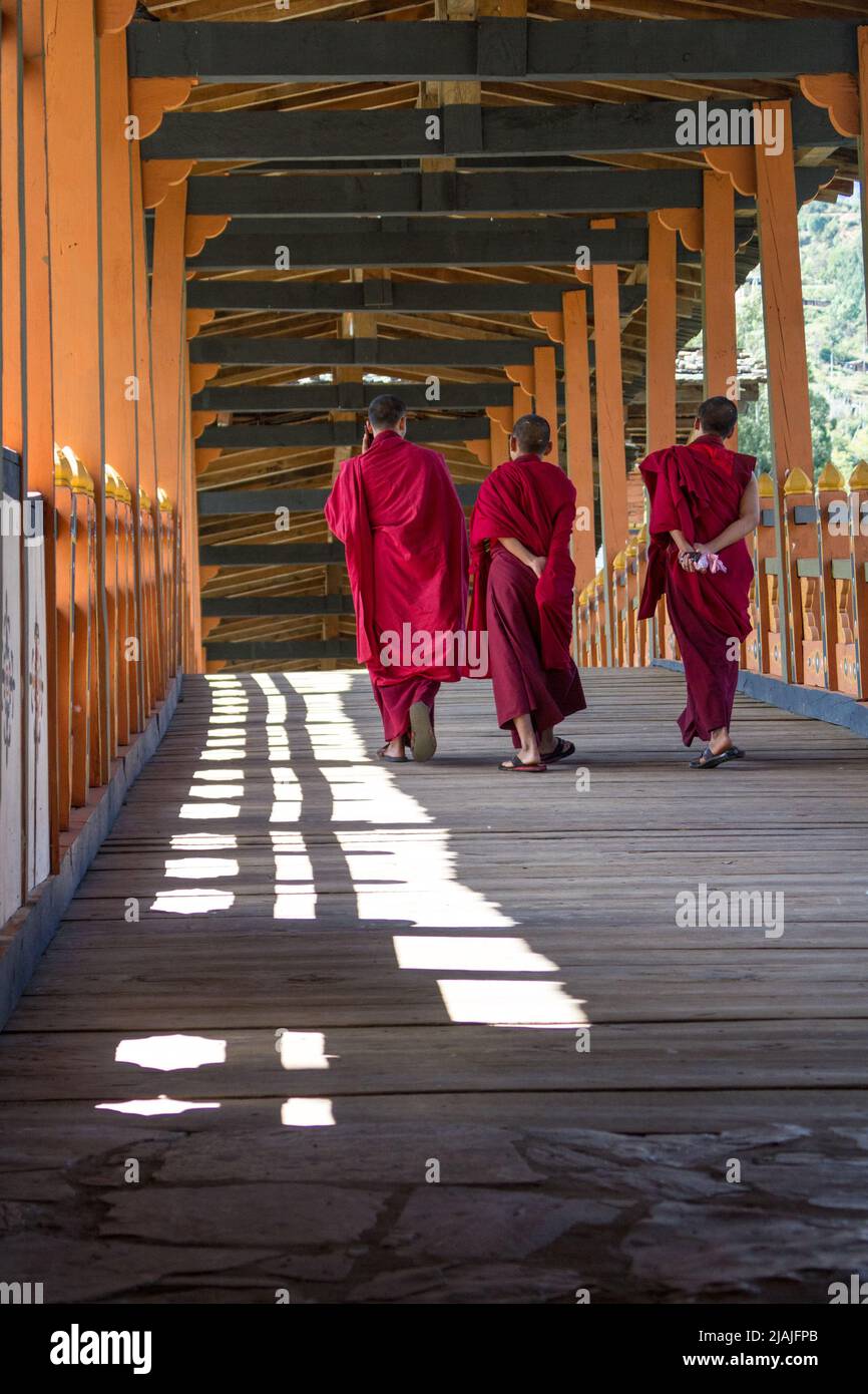 A group of robed monks walks over a covered bridge in Punakha, Bhutan, while one of the three talks on a cellphone. Stock Photo