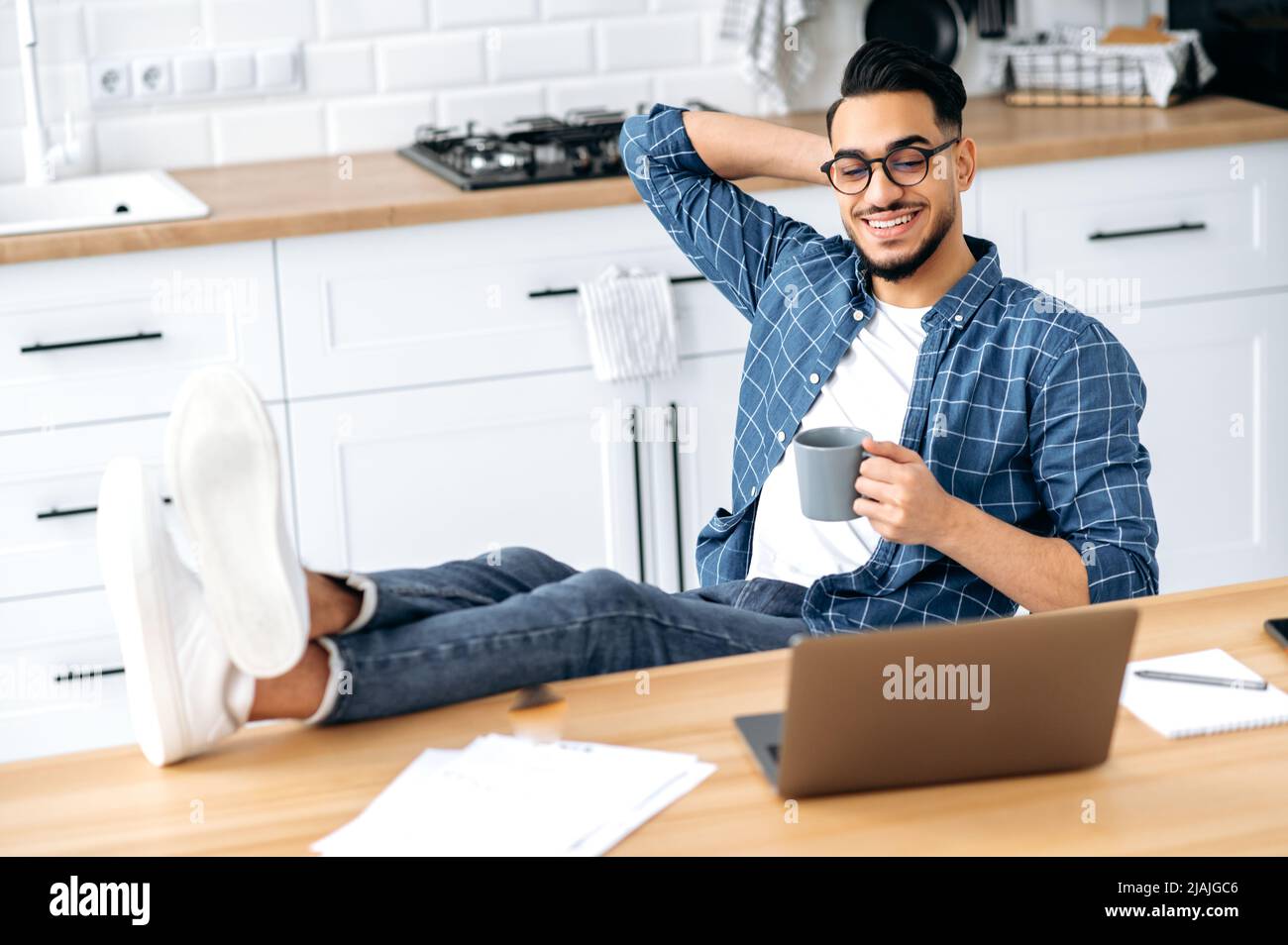 Chilling during work. Positive indian or arabian guy in stylish clothes, sit at home in the kitchen with his legs on the table, holding a cup of coffee in hand, working remotely from home using laptop Stock Photo