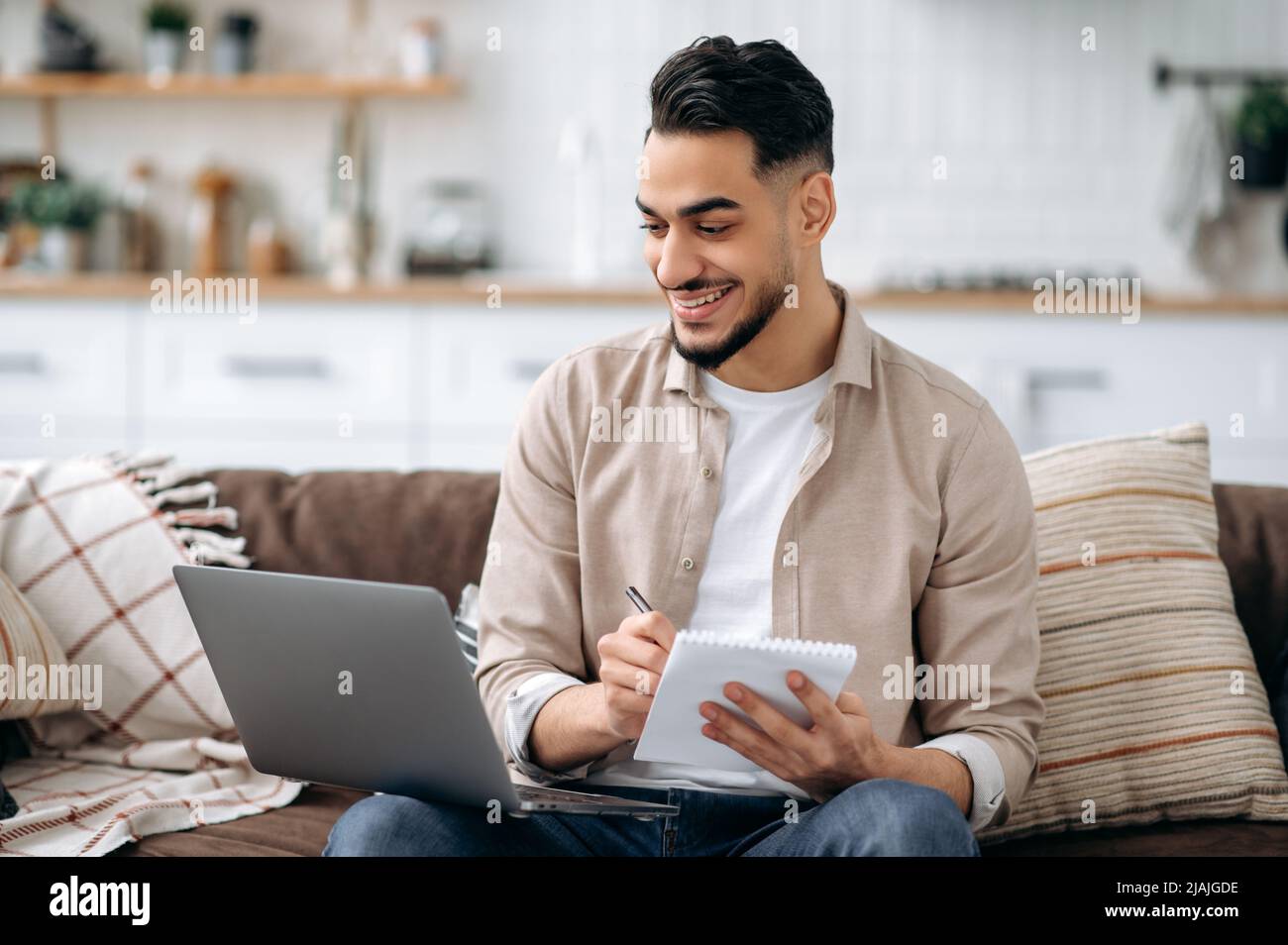 Positive smart indian or arabian man taking notes while working on project or learning online uses laptop, sitting on a sofa in a living room, browsing information in internet, prepares a report Stock Photo