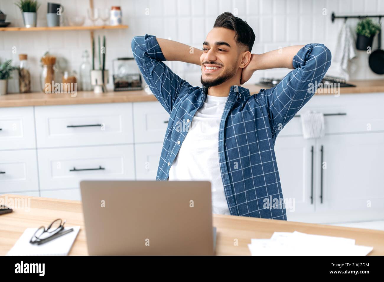 Happy arabian or indian guy, freelancer, working from home, sits in the kitchen at the workplace, takes a break from work, puts his hands behind his head, looks to the side, dreams of rest, smiles Stock Photo