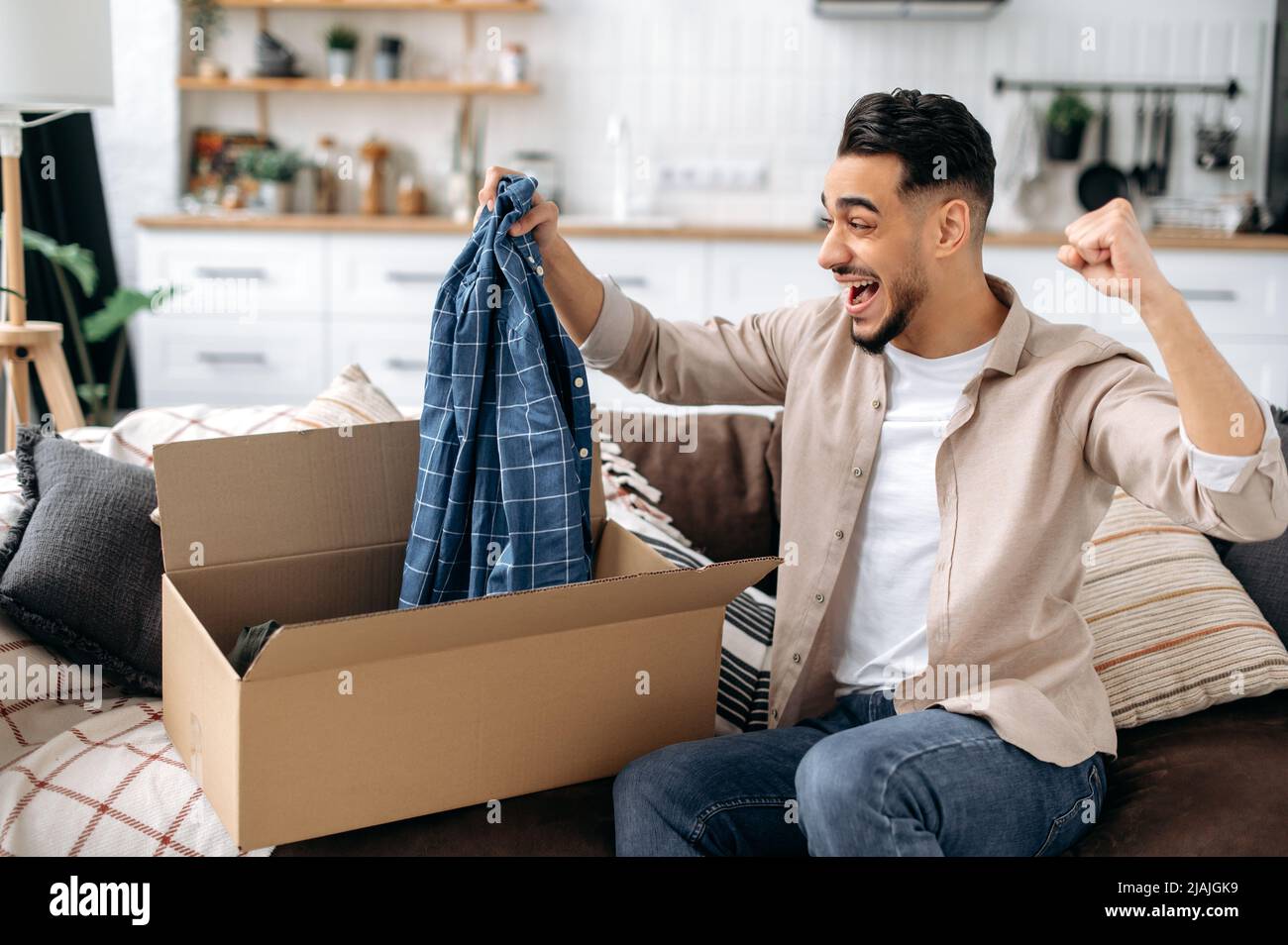 Happy attractive indian or arabian guy opening big carton box, unpacking internet store order at home, contented with fast delivery service and the goods which received, smiling and feel satisfied Stock Photo