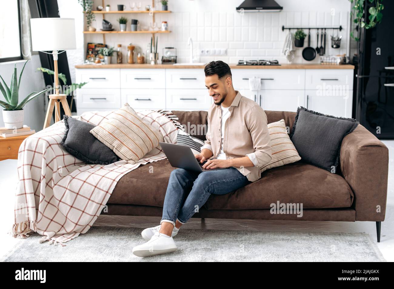 Positive handsome smart indian or arabian guy, freelancer or student, work or study online from home, using laptop, while sit on comfortable sofa in cozy living room, browsing internet, news, smiling Stock Photo