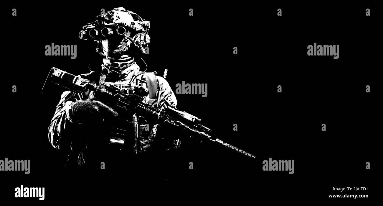Modern combatant standing in darkness, wearing night vision device and armed with silencer equipped assault rifle. Stock Photo