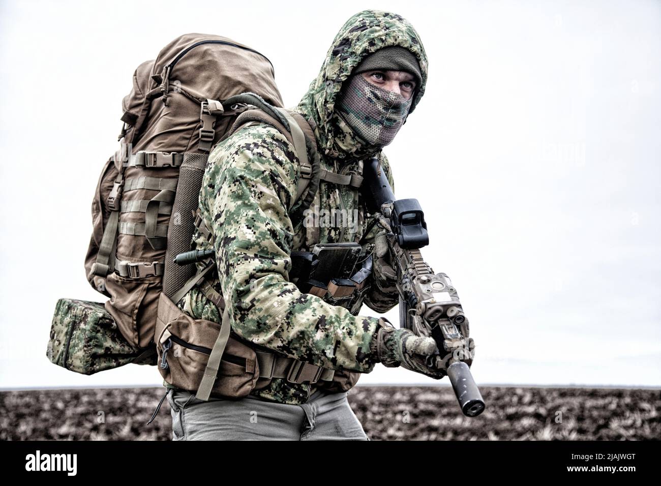 Military mercenary wearing hooded camo jacket and backpack, walking through muddy terrain. Stock Photo