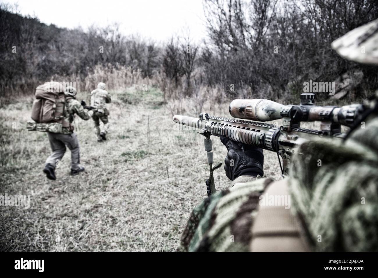 Over the shoulder view of a marksman covering comrades rushing through the woodlands. Stock Photo