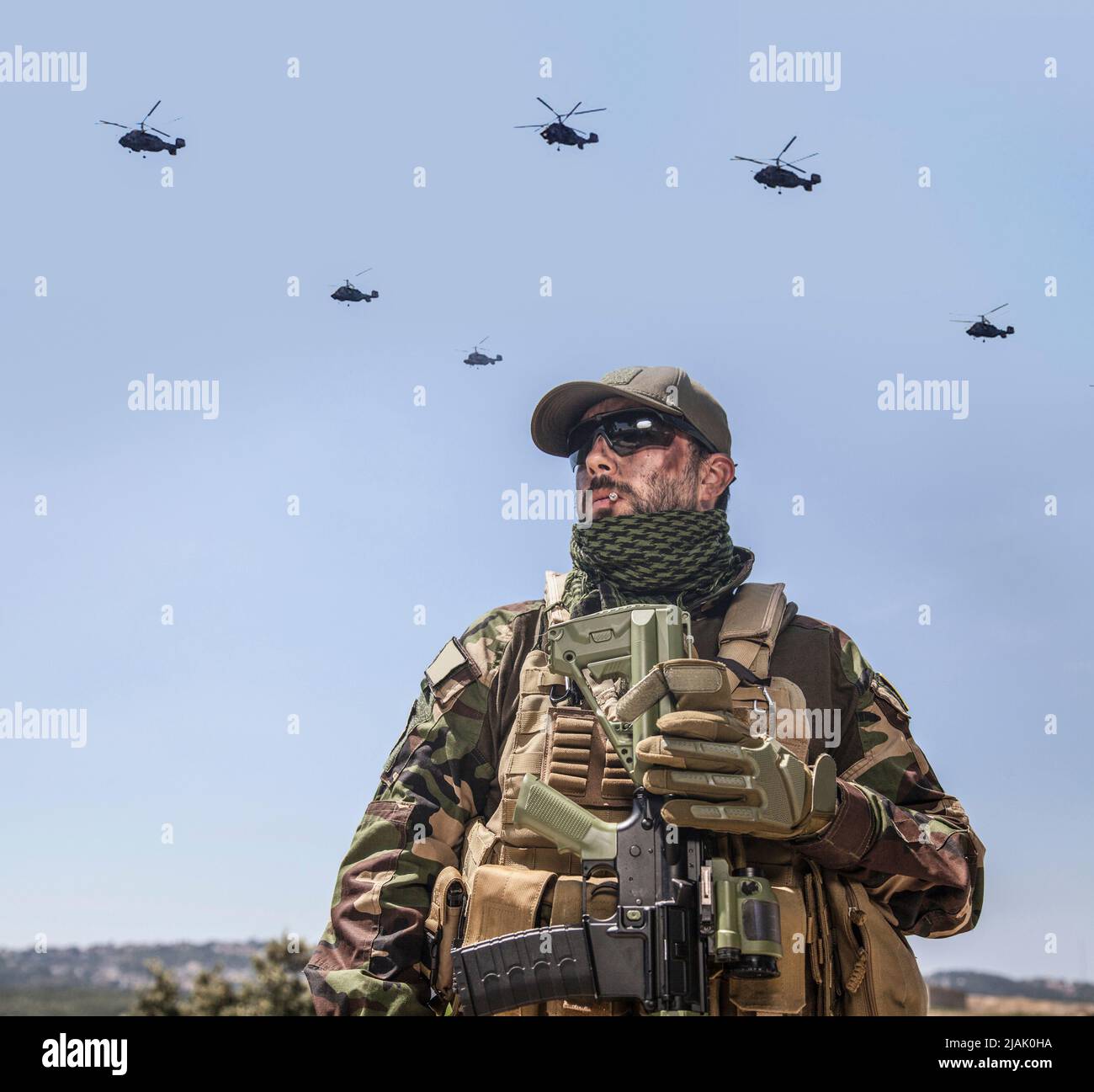 Soldier smoking a cigarette on ground while a convoy of helicopters fly overhead. Stock Photo