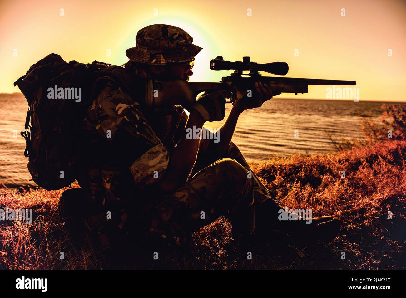 Silhouette of a sniper aiming rifle while sitting on the ocean shore during sunset. Stock Photo