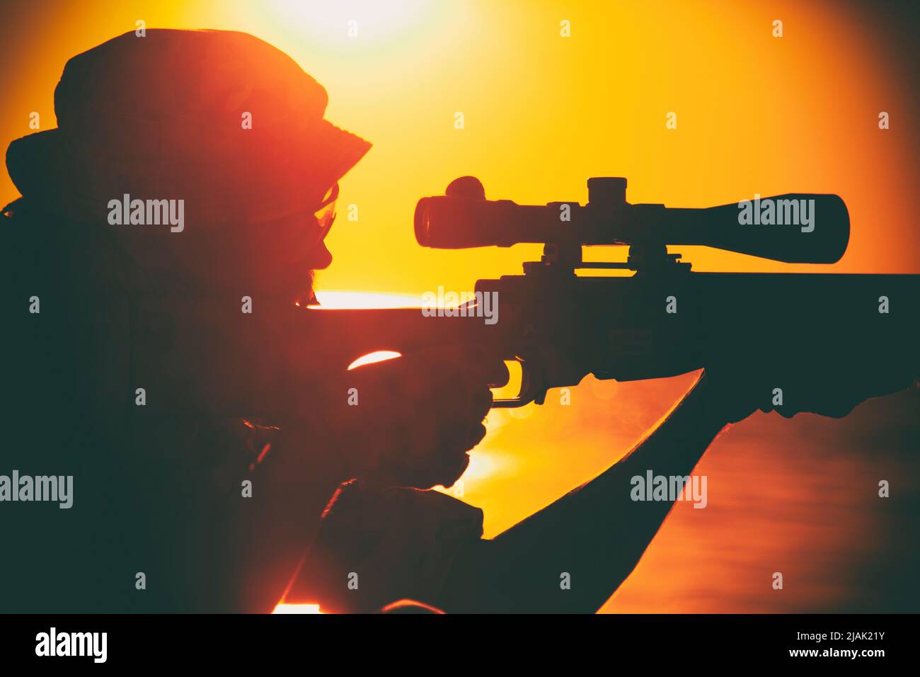 Silhouette of a sniper aiming rifle while sitting on the ocean shore during sunset. Stock Photo