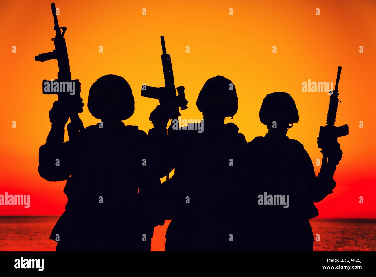 Silhouette of a group of soliders standing on shoreline at sunset, with assault rifles raised. Stock Photo