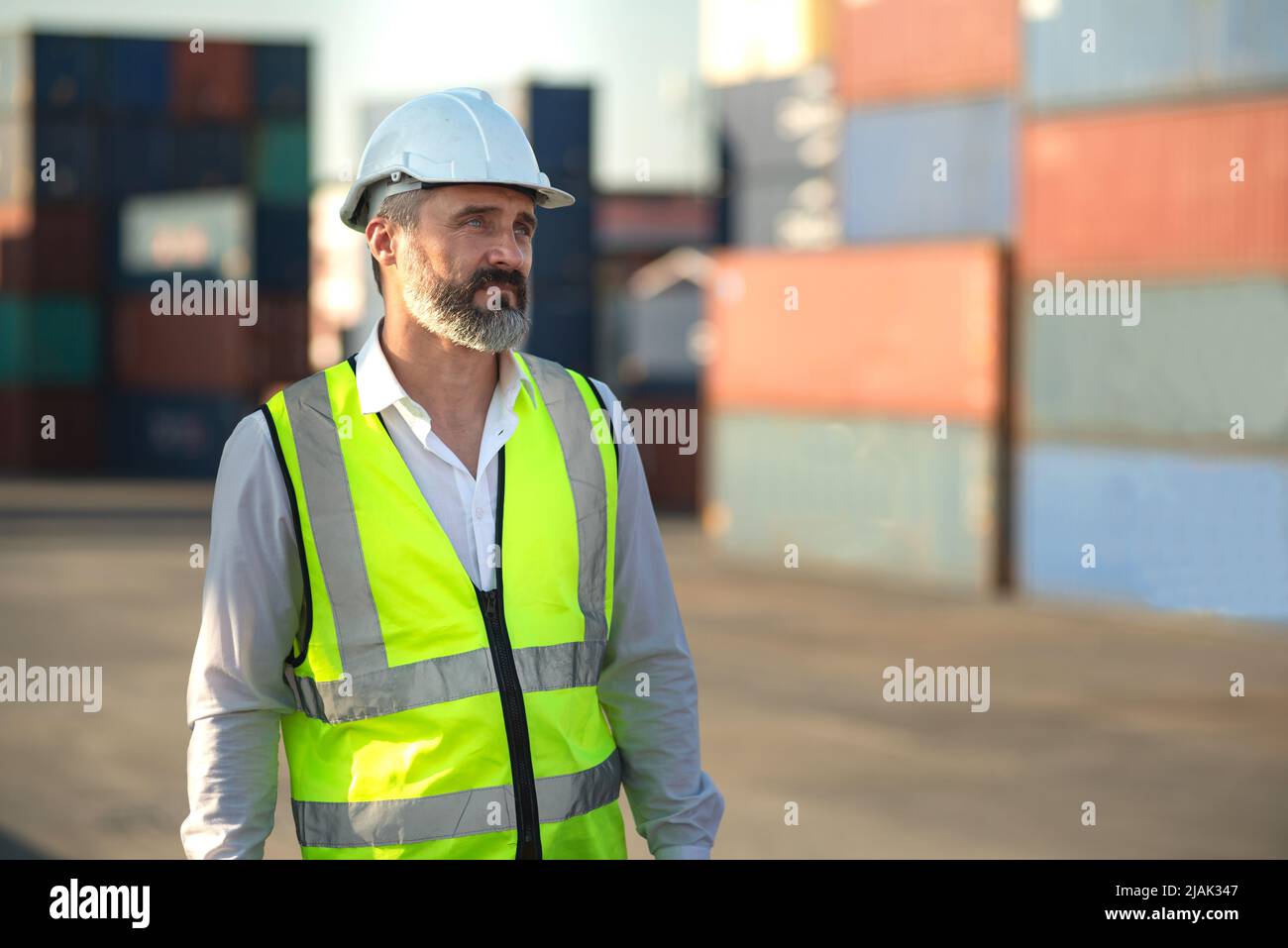 Caucasian male manager in white hard hat helmet and high-visibility vest walking in Container Terminal. Stock Photo