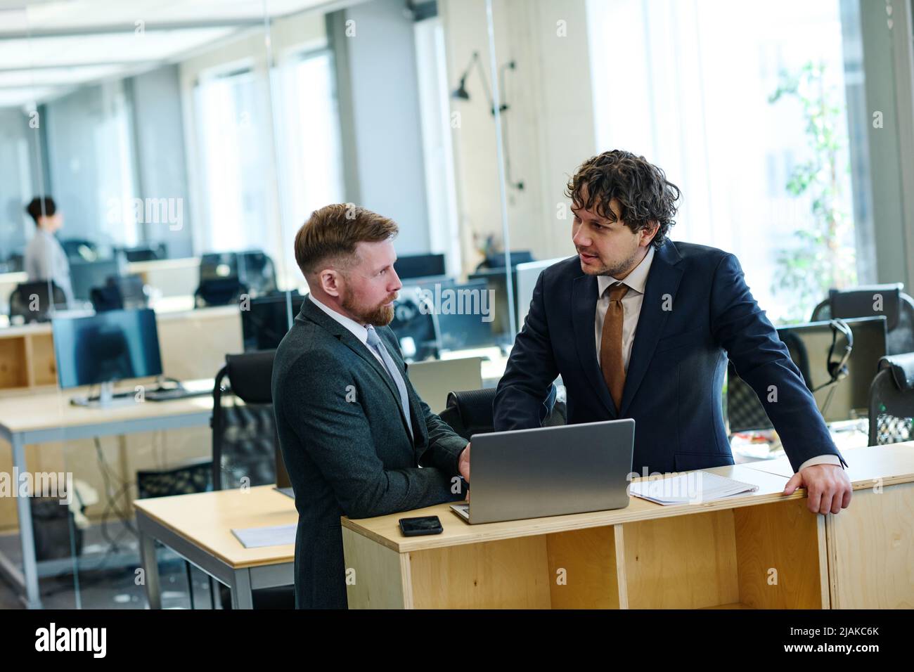 Two colleagues in formal wear standing at table at office and talking while using laptop Stock Photo