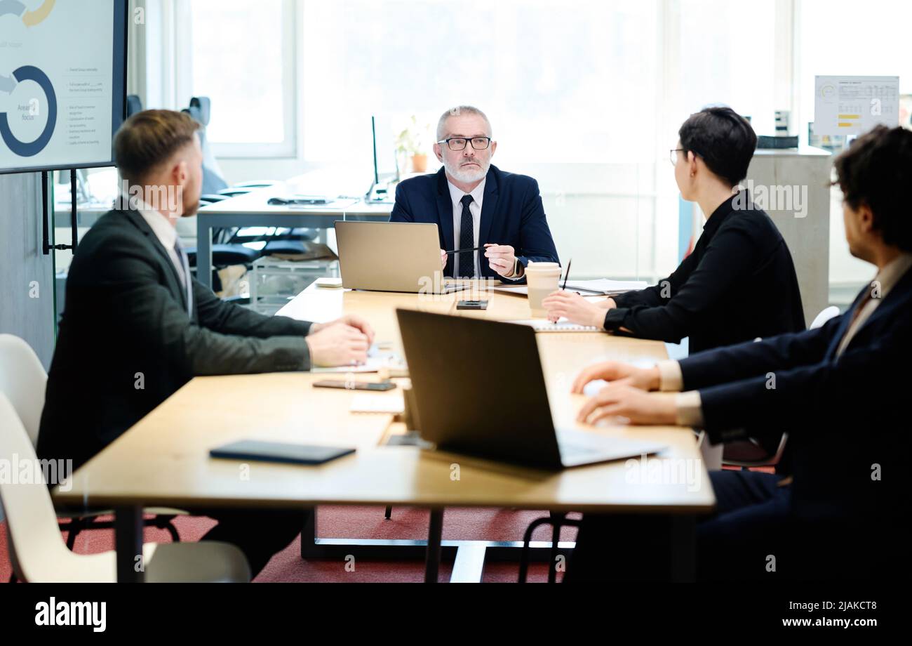 Senior manager in suit talking to partners at table with laptops during meeting at meeting room Stock Photo