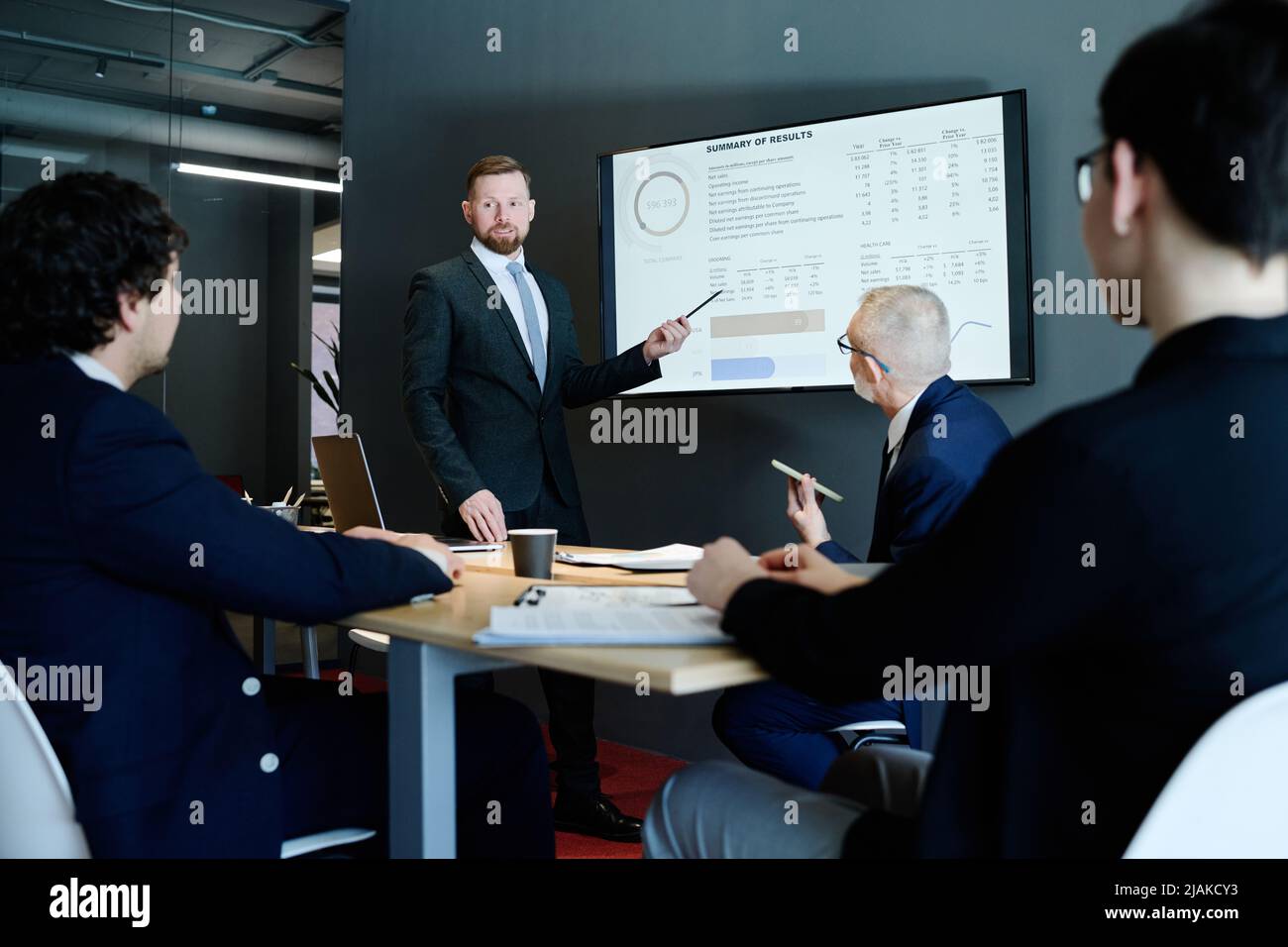 Young businessman in suit pointing at display screen with charts and performing with his presentation for colleagues at meeting room Stock Photo