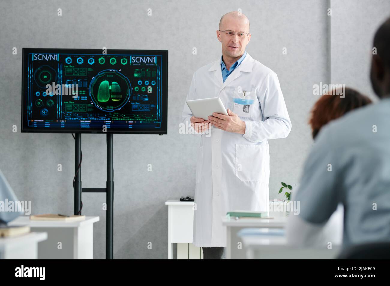 Mature lecturer in white coat with digital tablet standing near the display with human lungs and teaching students at class Stock Photo
