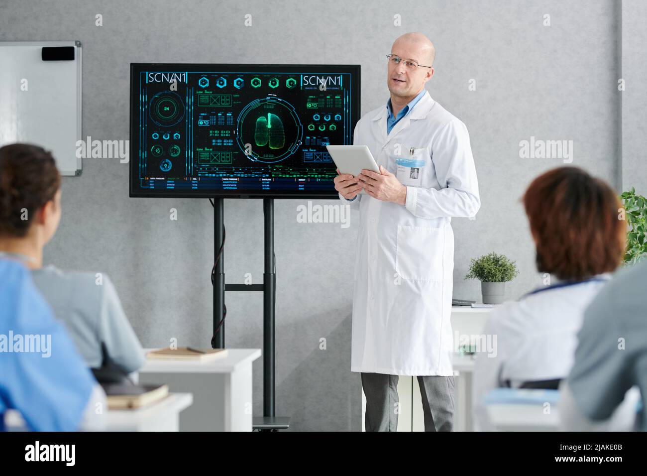 Mature lecturer in white coat standing at big display with medical charts on it and giving lecture to students Stock Photo