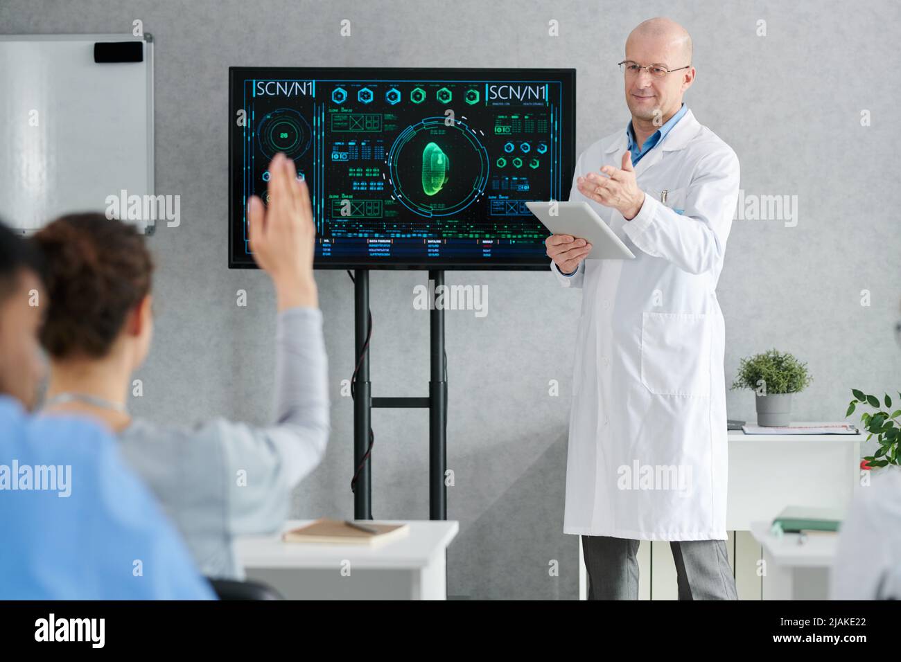 Mature doctor in lab coat with tablet pc asking question and pointing at girl who raising her hand at desk during seminar Stock Photo