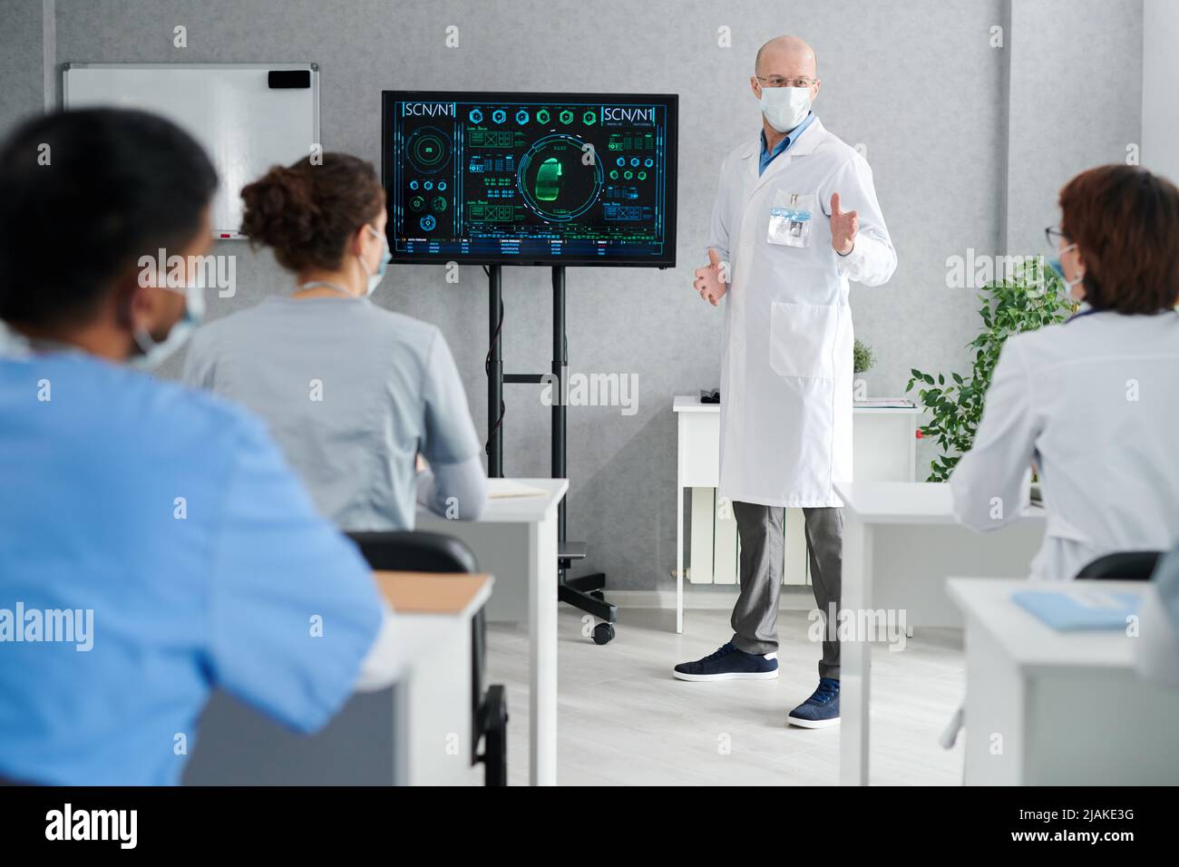 Professor in white coat and mask giving lecture about medical innovation for interns at desks at class Stock Photo