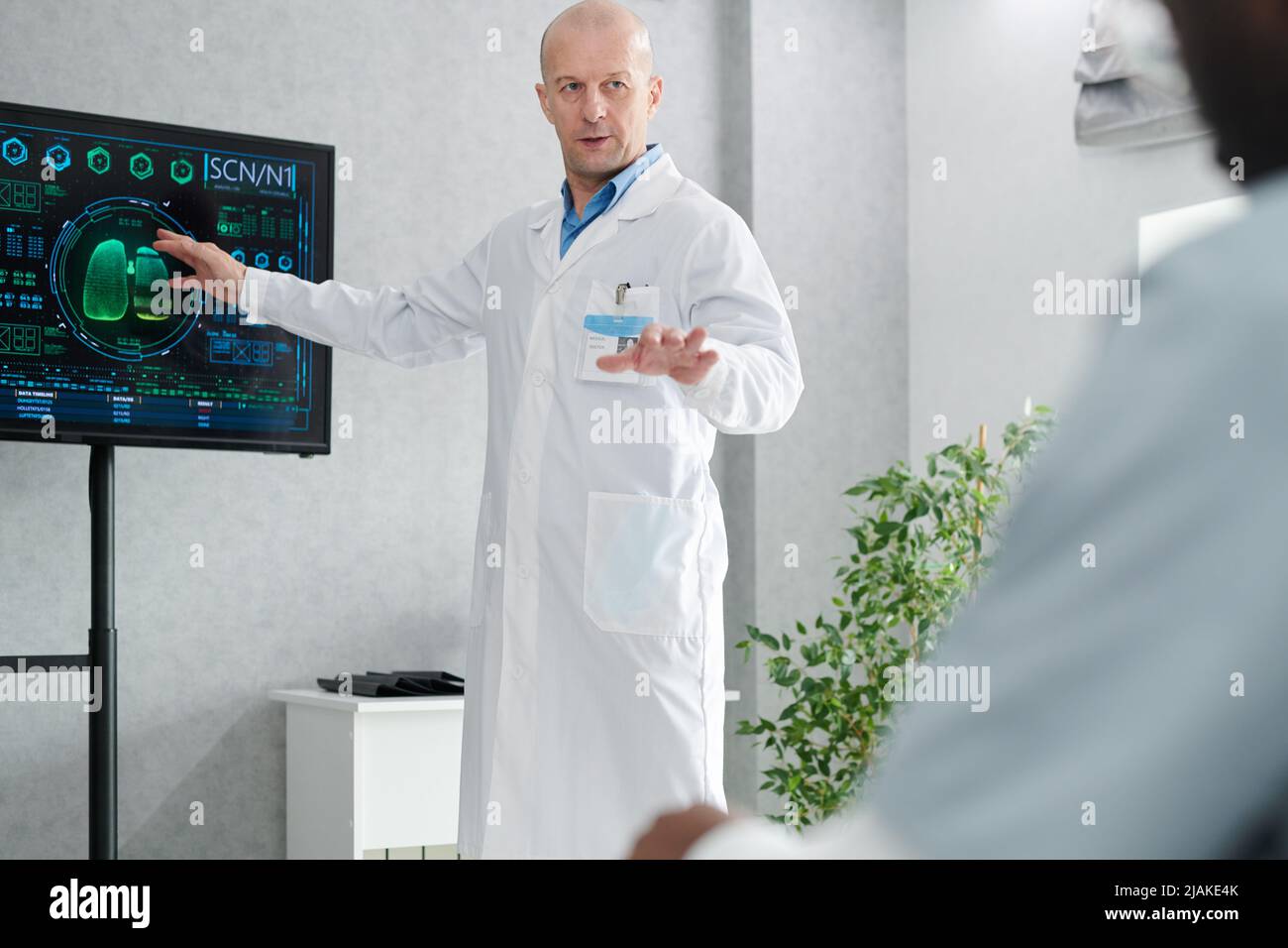 Mature doctor in white coat pointing at digital display with charts and talking to students during training Stock Photo