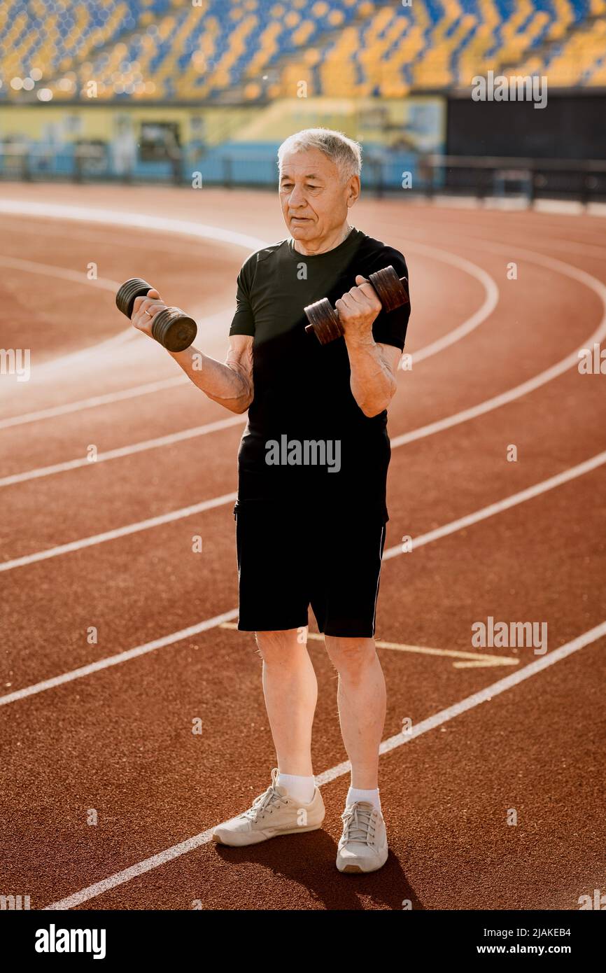 Senior man doing exercise with dumbbells in black sportswear outside. Mature concentrated man doing sports to keep fit. Stock Photo