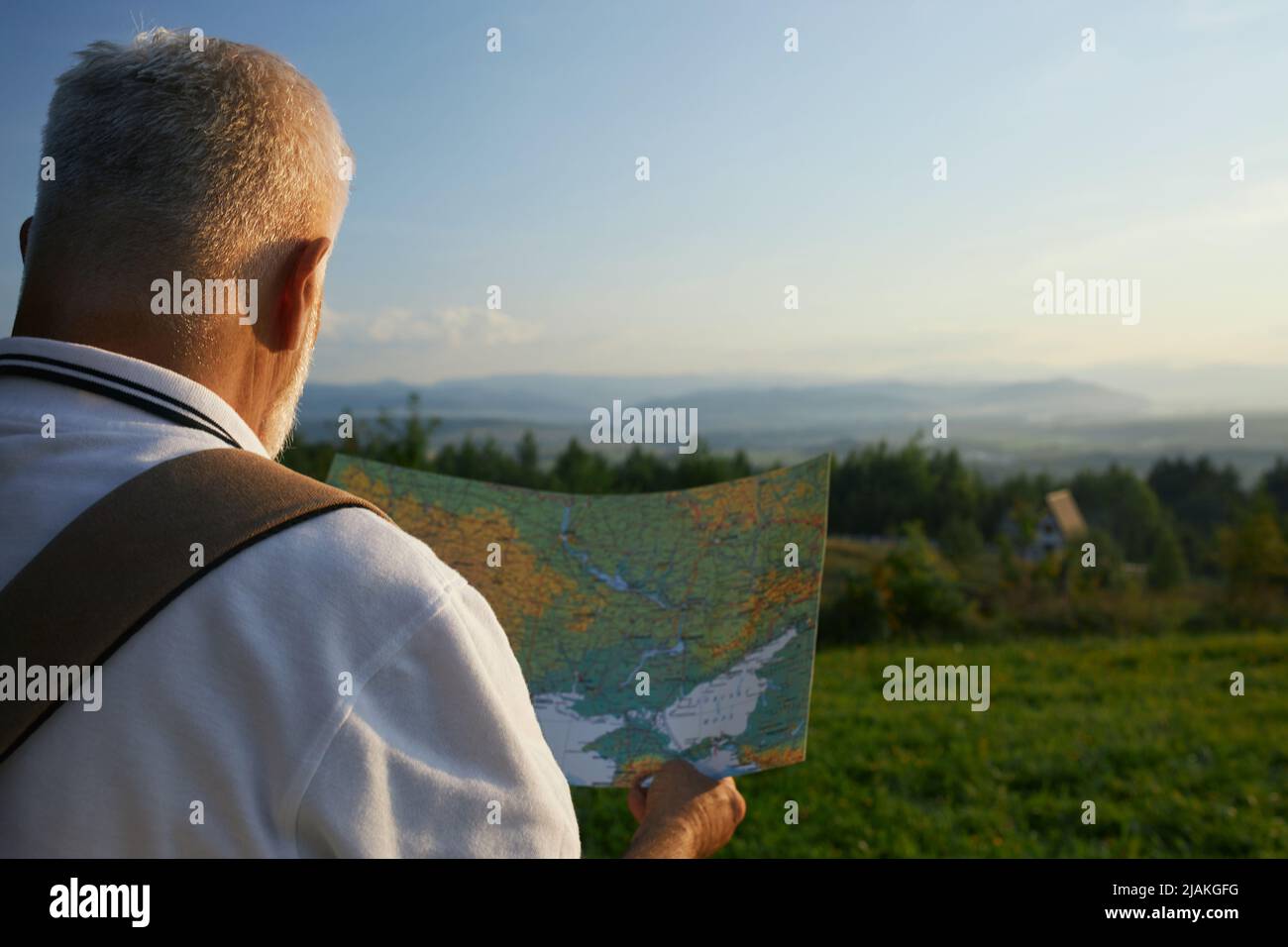 Adult male hiker checking touristic map, during mountain trip in summer. Back view of retired man holding map, looking for route, while hiking, with scenic landscape on background. Concept of tourism. Stock Photo