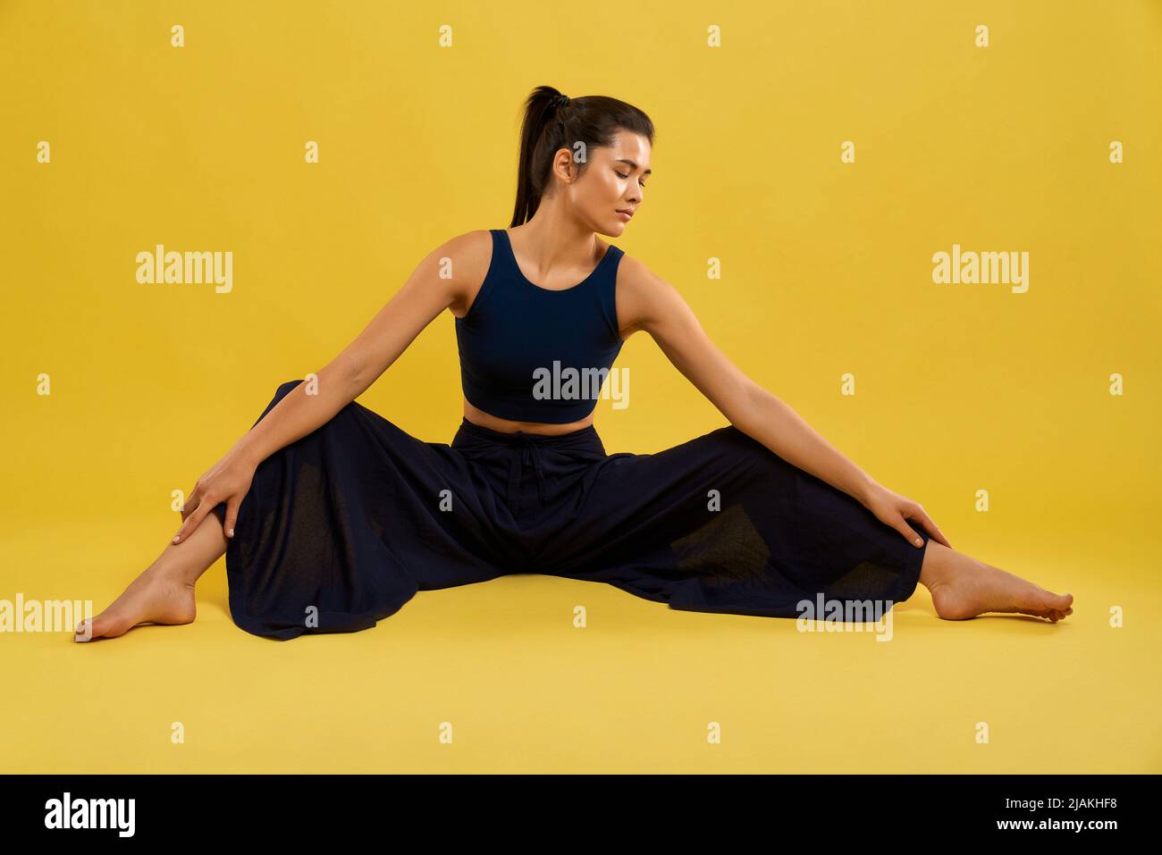 Slim young female yogi doing stretching exercise, sitting with legs wide apart indoors. Front view of pretty brunette girl stretching legs, isolated on orange studio background. Concept of yoga. Stock Photo